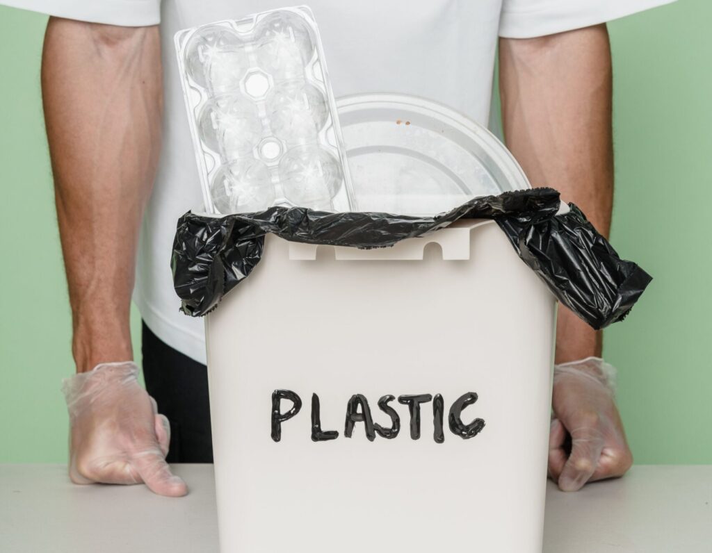 Adult male with gloves standing by a plastic recycling bin full of waste.
